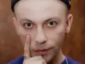 Zbliżenie na twarz Arka Kłusowskiego w czarnej czapce, z palcem przy policzku.