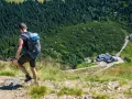 Mężczyzna z plecakiem wędruje po kamienistym szlaku w Karkonoszach, podziwiając widok na schronisko i las.