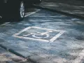 Niebieskie miejsce parkingowe dla niepełnosprawnych pod blokiem, z symbolem wózka inwalidzkiego. Obok widoczna część czarnego samochodu.