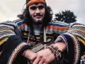 Młody mężczyzna w kolorowym swetrze w paski i opasce na głowie, uśmiecha się. Wygląda jak prawdziwy hippie.