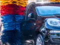 Czarna, błyszcząca karoseria samochodu w myjni automatycznej, otoczona kolorowymi szczotkami. Czy myjnia automatyczna niszczy lakier? Woda spływa po aucie.