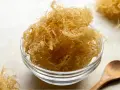 Un bol en verre rempli de **irish moss** séché, ressemblant à des fils fins et dorés. Une cuillère en bois repose à côté.
