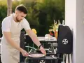 Mężczyzna w fartuchu przygotowuje mięso na grillu na balkonie. Czy można robić grilla na balkonie? Tak, jeśli jest bezpiecznie!