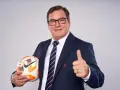 Uśmiechnięty trener reprezentacji Polski w garniturze trzyma piłkę i pokazuje kciuk w górę.