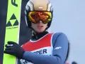 Młody skoczek narciarski w kasku i goglach, gotowy do skoku. Na nartach widnieje logo Fischer.
