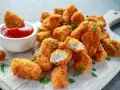 Chrupiące kawałki filet z kurczaka panierowany, jeden zanurzony w keczupie, na desce z ziołami.