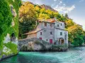 Antica casa in pietra con edera su un lago turchese, un ponte ad arco e colline verdi. Un luogo incantevole per i cammini in Lombardia.