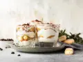 Pyszne desery włoskie: tiramisu w szklankach, udekorowane kakao i kawą. Obok kawałki biszkoptów i ziarna kawy.