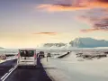 A silver van leads a convoy of vehicles driving in Iceland in December, past snow-covered mountains under a dramatic sunset sky.