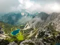 Blick vom Drachenkopf auf einen türkisfarbenen Bergsee, umgeben von grünen Hängen und felsigen Gipfeln, Wolken ziehen über die Landschaft.