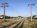 Linia średniego napięcia na działce, z domami w tle. Słupy energetyczne przecinają niebo, a ich cienie padają na suchą trawę.