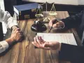 Prawnik zarządza masą upadłościową, omawiając dokumenty z klientem. Na stole młotek sędziowski, waga sprawiedliwości i księgi.
