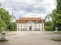 Pałac w Nieborowie, biała rezydencja z czerwonym dachem, otoczona zielenią. Dwie wieżyczki z zielonymi kopułami po bokach.