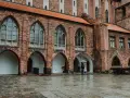 Zamek w Kwidzynie, z ceglanymi arkadami i gotyckimi oknami, skrywa tajemnice. Dwoje ludzi z parasolem spaceruje po mokrym dziedzińcu.