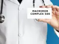 Lekarz w białym kitlu trzyma kartkę z napisem MACMIROR COMPLEX 500. Stetoskop wisi na jego szyi.