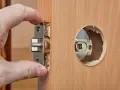 Dłoń montuje mechanizm zamka w drewnianych drzwiach. Dowiedz się, ile kosztuje wymiana zamka w drzwiach.