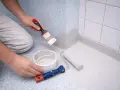 Person målar hörn på våtrumsmatta med pensel. En färgburk och en liten roller ligger bredvid.