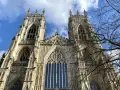 The majestic York Minster, a Gothic cathedral, stands tall against a bright blue sky with scattered clouds. Bare tree branches frame the intricate stonework and towering spires.