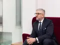 Mężczyzna w garniturze i okularach siedzi na czerwonym fotelu, patrząc w bok.