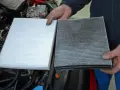 Hands hold a new white air filter next to a dirty, used one, ready to replace air filter in car.