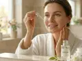 Kobieta w białym szlafroku pokazuje, jak nakładać serum na twarz. Na stole stoją butelka z serum i słoik z kremem.