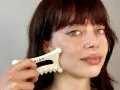 A woman with bangs and green eyes performs a facial fascia massage using a white gua sha tool.