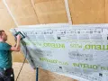 Mann bringt mit einem Tacker eine Dampfbremse an der Dachschräge an. Die Folie ist mit "INTELLO PLUS" bedruckt.