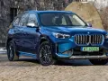 Niebieskie BMW iX1 stoi na kamienistej nawierzchni przed kamienną ścianą.