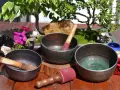 Tres cuencos tibetanos para limpiar piedras, con mazos de madera y detalles ornamentales, junto a un bonsái y flores.