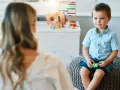 Chłopiec siedzi na pufie naprzeciwko psychoterapeutki, w tle zabawki.