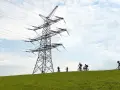 Grupa rowerzystów przemierza zielone wzgórze pod potężną konstrukcją linii 110 kV.