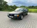 Ciemny Audi 80, klasyk z lat 90., idealny dla szukających aut do 5 tys. zł. Zaparkowany na kostce brukowej, otoczony zielenią.