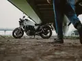 Biały motocykl klasyczny, kategoria A2, pod mostem. Obok przechodzi motocyklista z kaskiem.