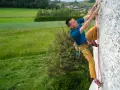 Mężczyzna uprawia wspinaczkę sportową na skalnej ścianie, z widokiem na zielone pola i wieś.