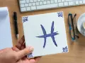 Dłoń trzyma kwadratową płytkę z symbolem ryby, znakiem zodiaku. Na biurku leżą notatnik, długopis, okulary i kawa.