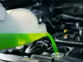 A gloved hand pours green antifreeze and coolant into a car's engine.