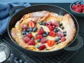 Ein goldbrauner Dutch Baby mit Puderzucker und frischen Beeren, serviert in einer gusseisernen Pfanne.
