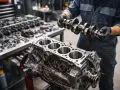 Mechanik w czarnych rękawiczkach trzyma wałek rozrządu nad blokiem silnika. Na stole leżą narzędzia i części, gotowe do remontu silnika 2T.