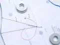 Sur un patron de couture, une aiguille rouge traverse le papier, retenue par des rondelles métalliques.