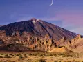 Krajobraz w parku narodowym Teide z księżycem w kształcie sierpa na fioletowym niebie. Wulkan góruje nad skalistym terenem.
