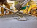 Pracownik wylewa beton, który będzie schnął. W tle koparka i budowa.