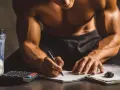 Un athlète calcule combien de protéine par jour il doit consommer, avec un shaker de poudre, une calculatrice et un carnet.