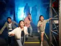 Grupa przerażonych ludzi ucieka przed postaciami w dymie. To wygląda jak prawdziwy escape room, gdzie trzeba rozwiązać zagadki, by przeżyć.