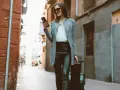 Kobieta w skórzanych spodniach i eleganckim płaszczu, z telefonem w ręku, ciągnie walizkę. Idealny przykład, co założyć do skórzanych spodni na podróż.