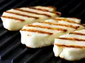 Trzy kawałki halloumi z grilla, z apetycznymi paskami, leżą na czarnej grillowej płycie.