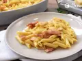 Plato de macarrones a la carbonara cremosos con trozos de panceta. En el fondo, un bol con más pasta y un rallador de queso.