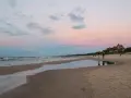 Zachód słońca nad plażą w Łebie. W oddali widać charakterystyczne budynki, a na piasku stoją spacerowicze.