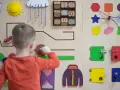 Chłopiec bawi się na tablicy edukacyjnej, rozwijając umiejętności manualne zgodnie z metodą Montessori. Widoczne są kolorowe kształty, zębatki i zamek.