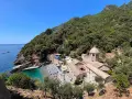 Baia con spiaggia affollata e un antico monastero, un luogo ideale per i cammini in Liguria, con mare azzurro e vegetazione rigogliosa.