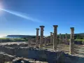 Sunlight streams over ancient roman ruins in Spain, illuminating weathered stone columns and walls against a bright blue sky and distant coastline.